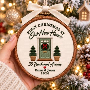 May include: A round wooden embroidery hoop ornament featuring white fabric with stitched text First Christmas at Our New Home, 35 Beechwood Avenue, Emma & James, 2026, a green front door with a wreath, two potted trees, and a cream ribbon bow.