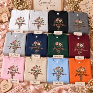 May include: A collection of twelve crewneck sweatshirts in various colors, including white, ash, black, grey, sport grey, navy, forest green, maroon, light pink, sand, light blue, and orange. Each sweatshirt features an embroidered floral bouquet with the word grandma in cursive script.