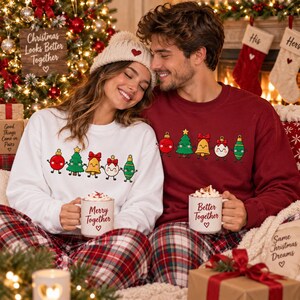 May include: A white sweatshirt with a graphic of five cartoon Christmas ornaments holding hands and a matching maroon sweatshirt with the same graphic. Both sweatshirts are paired with red and white plaid pants and white ceramic mugs.