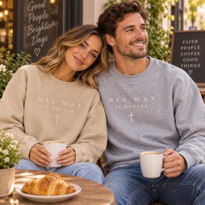 May include: A beige crewneck sweatshirt and a grey crewneck sweatshirt, both featuring the text "HIS WAY IS BETTER" printed in white above a small white cross symbol. The garments have long sleeves, ribbed cuffs, and a classic round neckline.