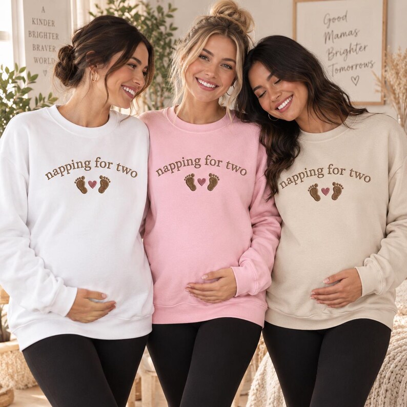 May include: Three crewneck sweatshirts in white, light pink, and beige, each featuring the brown text napping for two, two small brown baby footprints, and a small pink heart centered on the chest area above the waistline.