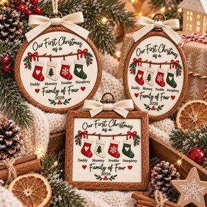 May include: Three wooden-framed ornaments featuring embroidered text Our First Christmas as a Family of Four 2026. The designs include four hanging stockings in red and green with white accents, topped with a cream-colored fabric ribbon bow.