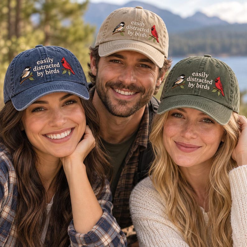 May include: Three baseball caps in navy blue, tan, and olive green. Each cap features the text "easily distracted by birds" embroidered in white, accompanied by a small illustration of a black-and-white bird and a bright red cardinal on branches.