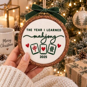 May include: A round wooden embroidery hoop ornament featuring white fabric with green embroidered text that reads THE YEAR I LEARNED mahjong and 2025. Three mahjong tiles are stitched below with red hearts, topped with a dark green velvet bow.