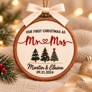 May include: A round wooden embroidery hoop ornament featuring cream fabric with embroidered text reading OUR FIRST CHRISTMAS AS Mr & Mrs, Martin & Elaine, and 09.21.2024. Three embroidered pine trees sit above the names, topped with a cream ribbon bow.