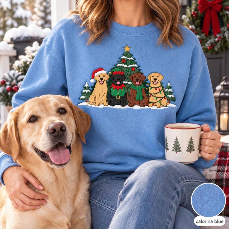 May include: A light blue crewneck sweatshirt featuring an embroidered graphic of four dogs sitting in front of a Christmas tree. The dogs include a yellow Labrador in a Santa hat, a black Labrador in an elf outfit, a brown Labrador, and a yellow Labrador.