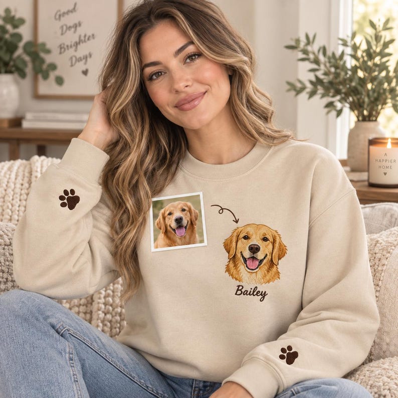 May include: A beige crewneck sweatshirt featuring a custom embroidered illustration of a golden retriever dog with the name Bailey written below it. The design includes a small paw print on each sleeve cuff and a square photo of the dog.