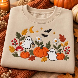 May include: A beige crewneck sweatshirt featuring an embroidered design of two white ghosts, orange pumpkins, red berries, a red and white mushroom, green leaves, white daisies, a yellow crescent moon, small stars, and two black bats.