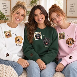 May include: Three crewneck sweatshirts in white, dark green, and light pink. Each features a small square dog photo, a matching illustrated dog portrait with a name below, and a small embroidered paw print on the sleeve cuff.