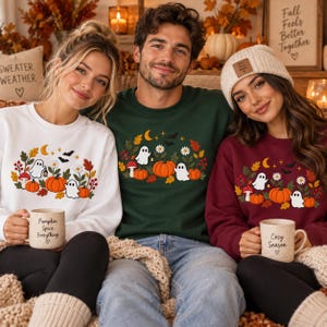 May include: Three crewneck sweatshirts in white, dark green, and maroon, each featuring a graphic of ghosts, pumpkins, autumn leaves, flowers, and bats. Two people hold ceramic mugs with the text Pumpkin Spice Everything and Cozy Season.