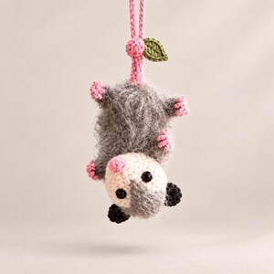 May include: A crocheted opossum hanging upside down by a pink yarn loop. The opossum has a fuzzy grey body, a cream-colored face with black eyes, a pink nose, small pink paws, and black ears. A small green crocheted leaf is attached to the pink loop.