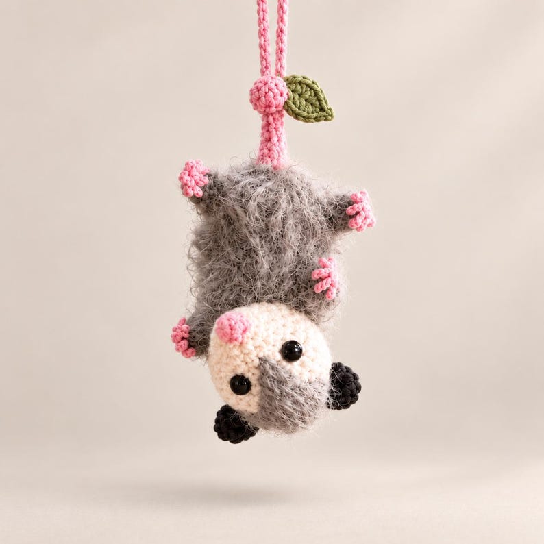 May include: A crocheted opossum hanging upside down by a pink yarn loop. The opossum has a fuzzy grey body, a cream-colored face with black eyes, a pink nose, small pink paws, and black ears. A small green crocheted leaf is attached to the pink loop.