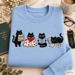 May include: A light blue crewneck sweatshirt featuring an embroidered graphic of four black cats. One cat holds a stack of books, one sits under a heart-patterned blanket reading, one wears glasses while reading, and one lies by a mug and tablet.