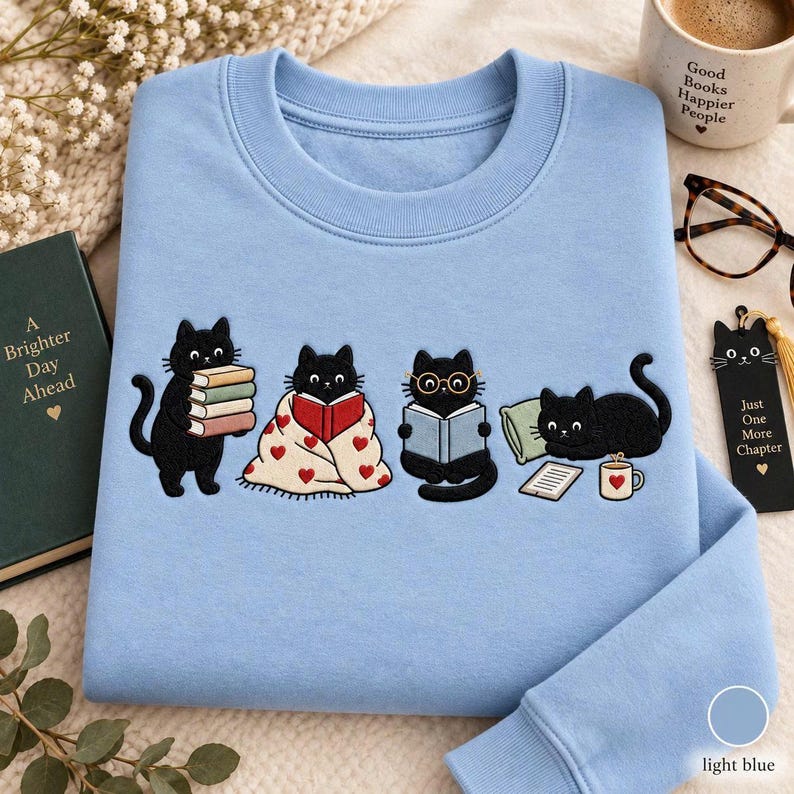 May include: A light blue crewneck sweatshirt featuring an embroidered graphic of four black cats. One cat holds a stack of books, one sits under a heart-patterned blanket reading, one wears glasses while reading, and one lies by a mug and tablet.