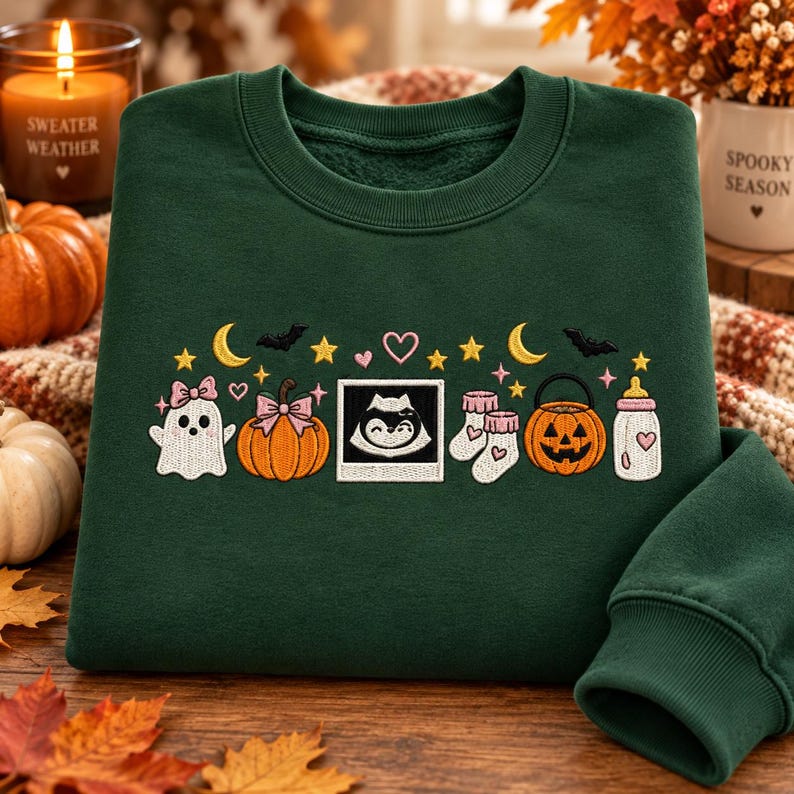 May include: A dark green crewneck sweatshirt featuring embroidered Halloween-themed icons including a ghost with a pink bow, a pumpkin with a pink bow, a sonogram image, baby socks, a jack-o'-lantern, and a baby bottle, accented by stars and moons.