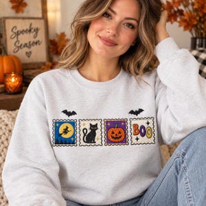 May include: A light gray crewneck sweatshirt featuring four embroidered rectangular patches arranged in a row. The patches depict a witch on a broomstick against a yellow moon, a black cat, a jack-o'-lantern, and the colorful text BOO.