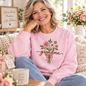 May include: A light pink crewneck sweatshirt featuring an embroidered bouquet of colorful flowers tied with a pink ribbon and the word grandma written in brown cursive script across the center of the chest area of the garment.