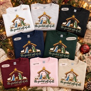 May include: A collection of eight crewneck sweatshirts in white, ash, black, navy, forest green, maroon, light pink, and sand. Each features a nativity scene graphic with the text "the greatest gift of all" and "Matthew 1:21" printed below.