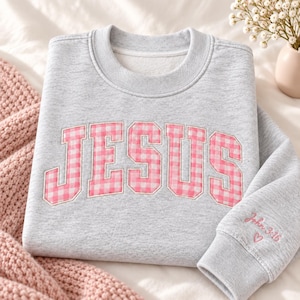 May include: A heather gray crewneck sweatshirt featuring the word JESUS in large, pink and white gingham patterned block letters across the chest. The right sleeve cuff is embroidered with the text John 3:16 and a small pink heart outline.