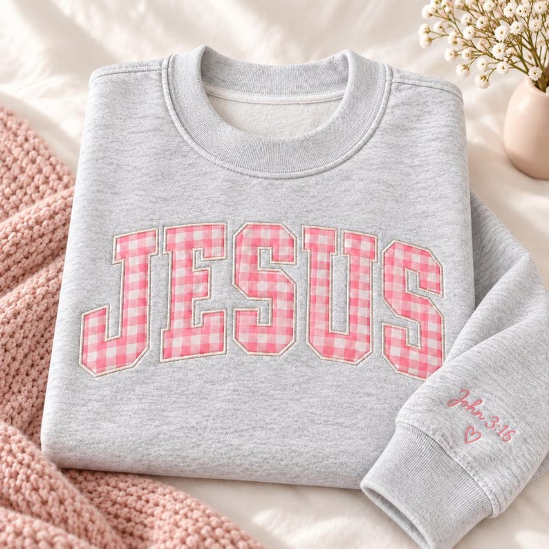 May include: A heather gray crewneck sweatshirt featuring the word JESUS in large, pink and white gingham patterned block letters across the chest. The right sleeve cuff is embroidered with the text John 3:16 and a small pink heart outline.