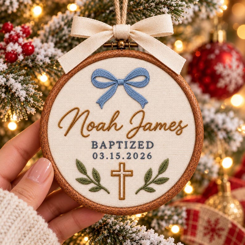 May include: A round wooden embroidery hoop ornament featuring cream fabric with a blue embroidered bow, the name Noah James in brown script, the text BAPTIZED 03.15.2026, a gold cross, and green leaves, topped with a cream fabric bow.