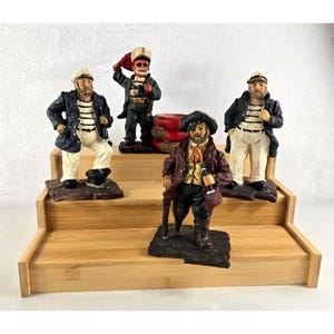 Op de afbeelding: Vier nautische beeldjes op een houten display. De beeldjes beelden zeelieden en een piraat uit, die elk voorwerpen vasthouden zoals flessen en een wandelstok. De figuren dragen hoeden, jassen en gestreepte shirts. Het kleurenpalet omvat zwart, wit, bruin en rood.