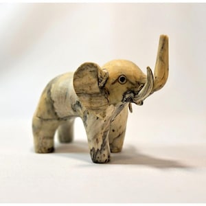 May include: A small, carved elephant figurine with a trunk raised upwards. The elephant is cream-colored with dark gray marbling throughout its body. It has large ears, small tusks, and a detailed eye. The figurine is standing on a white surface.