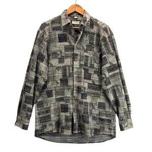 Puede incluir: Una camisa abotonada de manga larga con un estampado geométrico en tonos grises y negros. La camisa está colgada de una percha de madera. La etiqueta dice "GIORGIO ARMANI".