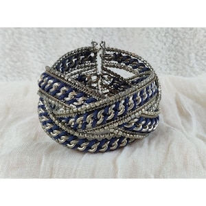 Op de afbeelding: Een geweven zilveren en blauwe armband met meerdere lagen ketting en koord.