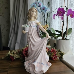 Könnte beinhalten: Eine Porzellanfigur einer Person mit langem blondem Haar, die ein fließendes, blassrosa Kleid mit einem blauen Taillenband trägt. Die Figur hält einen kleinen weißen Topf mit hellblauen Blumen und hat aufgemalte rosa Rosen am Saum.