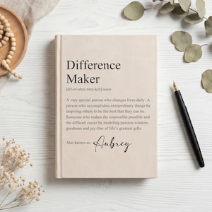 Puede incluir: Un diario de tapa dura en color beige que presenta la definición impresa de Difference Maker, incluyendo la ortografía fonética, un párrafo descriptivo y el nombre Aubrey en fuente cursiva, acompañado por una pluma estilográfica negra.