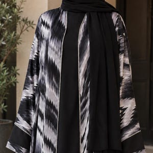 May include: A black and white open-front abaya featuring a bold, repeating chevron-inspired zigzag pattern in shades of gray. The garment is worn over a solid black dress and includes a matching black headscarf, with solid black cuffs at the sleeves.