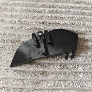 May include: A black 3D-printed plastic component with a curved, tapered edge and a flat top surface. It features a central rectangular protrusion with a small square cutout, flanked by two smaller vertical rectangular tabs and a notched end.