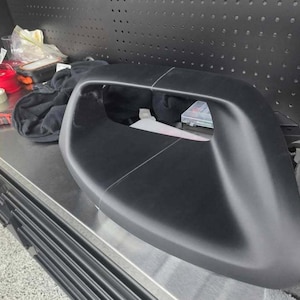 Op de afbeelding: Een matzwarte motorkap-luchthapper met een brede, gebogen inlaatopening en een centrale naadlijn die verticaal over het vooroppervlak loopt. Het onderdeel heeft een gladde, gevormde afwerking en een holle interne structuur.