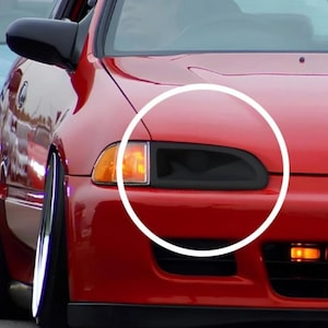 Op de afbeelding: Een felrode auto met een op maat gemaakte zwarte koplampafdekking. Het voertuig heeft een verlaagde stand met zichtbaar negatief camber op de voorwielen. Een oranje richtingaanwijzer bevindt zich naast de zwarte koplampbehuizing.