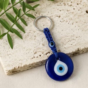 May include: A keychain featuring a circular dark blue glass evil eye charm with a white and light blue center. The charm is attached to a braided blue cord that includes a smaller matching evil eye bead and a silver-toned metal ring.