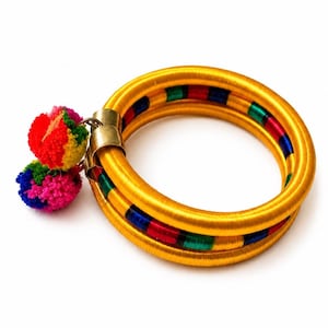 May include: A set of two circular bangles wrapped in bright yellow thread, featuring alternating segments of red, green, and blue thread. The bangles are connected by a gold-toned metal clasp adorned with two multi-colored yarn pom-poms.