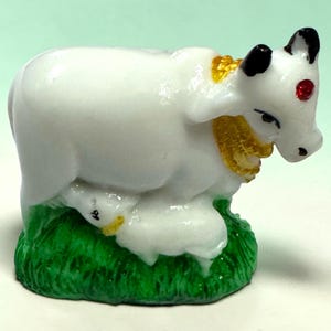 May include: A small white cow figurine with black-tipped horns and a red dot on its forehead. The cow wears a gold-coloured collar and stands on a textured green base, with a small white calf nestled at its side, also featuring yellow accents.