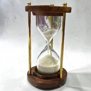 Puede incluir: Un reloj de arena con un bulbo de vidrio transparente lleno de arena blanca, sostenido por un marco de madera marrón oscuro y cuatro varillas metálicas delgadas de tono dorado. La estructura consta de una base circular de madera y una tapa a juego.
