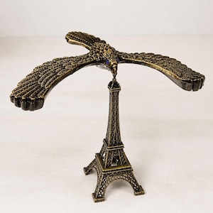 May include: A bronze-colored metal balancing eagle figurine with outstretched wings and small blue eyes, perched by its beak on the apex of a miniature Eiffel Tower structure. The entire object features a textured, antique-style metallic finish.