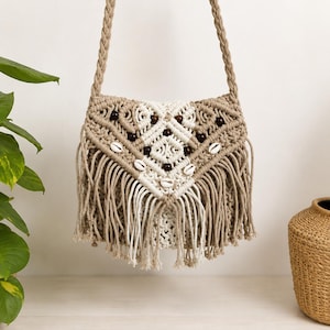 May include: A macrame shoulder bag crafted from tan and white cord, featuring a braided strap, intricate knot patterns, dark brown wooden beads, and white seashell accents. The front flap displays a V-shaped design with long, hanging fringe.