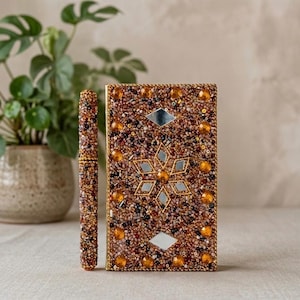 May include: A rectangular notebook and a matching cylindrical pen, both covered in a dense layer of brown, gold, and black beads. The notebook features a central star-shaped arrangement of small mirrors and additional amber-coloured cabochons.