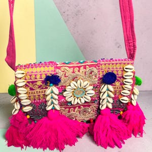 May include: A bright pink crossbody bag featuring intricate gold embroidery, a central flower motif made of white cowrie shells, and dangling tassels. The bag is adorned with additional cowrie shell accents, blue pom-poms, and green pom-poms.