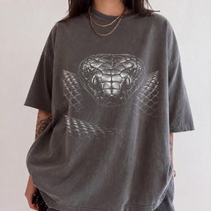 May include: A charcoal gray short-sleeve t-shirt featuring a large, detailed white graphic print of a snake head centered on the chest. The shirt has a crew neckline and a relaxed, loose fit, showcasing the reptile's scales and eyes in high contrast.
