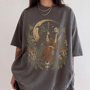 May include: A charcoal gray short-sleeve t-shirt featuring a graphic print of a figure in a pointed hat holding a glowing orb. The design includes a large crescent moon, stars, mushrooms, crystals, and foliage in muted gold, green, and orange tones.