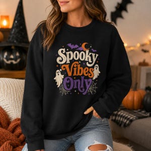 Puede incluir: Una sudadera negra de manga larga con un estampado gráfico que incluye el texto Spooky Vibes Only en blanco, naranja y morado. El diseño presenta ilustraciones de dos fantasmas blancos, murciélagos morados, una luna creciente y telarañas.