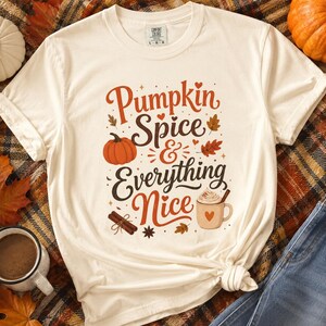 Puede incluir: Una camiseta de manga corta de color crema con el texto "Pumpkin Spice & Everything Nice" en letras marrones y naranjas. El gráfico incluye una pequeña calabaza naranja, una taza de café con nata y ramas de canela.