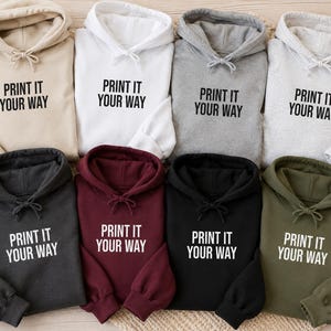 May include: A collection of eight hooded sweatshirts in various colors including beige, white, light gray, heather gray, dark gray, maroon, black, and olive green. Each hoodie features a drawstring hood and the text PRINT IT YOUR WAY.