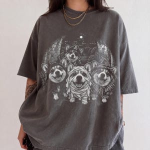 May include: A charcoal gray short-sleeve t-shirt featuring a graphic print of four Corgi dogs dressed in Halloween costumes, including a pirate, a devil, a mummy, and a scarecrow, set against a dark, circular background with brick details.