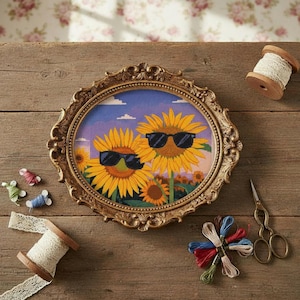 May include: An oval cross-stitch embroidery featuring two large yellow sunflowers wearing black sunglasses against a purple and blue sky, encased in an ornate gold frame. The piece is surrounded by spools of lace, embroidery thread, and metal scissors.